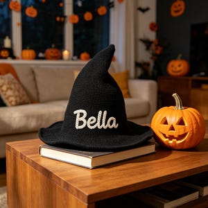 Puede incluir: Un sombrero de bruja de ganchillo negro con una corona puntiaguda y un ala ancha. La palabra Bella está bordada en letra cursiva blanca en la parte delantera del sombrero. El sombrero descansa sobre un libro cerrado junto a una calabaza.