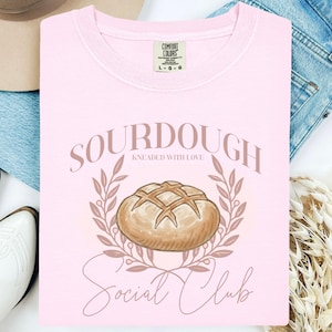 May include: A pale pink Comfort Colors sweatshirt featuring the words "SOURDOUGH KNEADED WITH LOVE Social Club" in brown script. The design includes a brown illustration of a bread loaf, encircled by a brown wreath.
