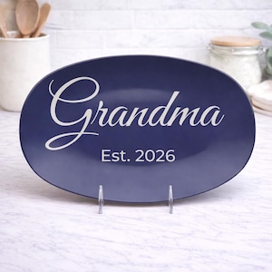 May include: A navy blue oval serving platter with the word "Grandma" in white script and the text "Est. 2026". The platter is displayed on a clear metal stand, perfect for serving food.