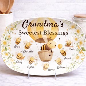 May include: Oval ceramic serving platter with the text "Grandma's Sweetest Blessings". The platter features a yellow hot air balloon with a bee in a basket, surrounded by cartoon bees and floral accents. Names are written below the bees.
