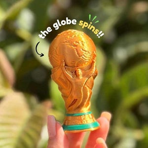 May include: A gold-colored trophy featuring a globe held aloft by figures, with a teal base and the text "WORLD CUP" printed on it. The text "the globe spins!!" is visible above the trophy, indicating the globe component rotates.