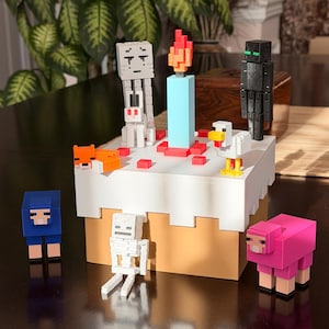 Könnte beinhalten: Eine Minecraft-Geburtstagstorte mit verschiedenen Charakterfiguren. Die Torte hat eine blaue Kerze mit einer roten und orangefarbenen Flamme und ist von Figuren wie einem Skelett, einem Schaf und einem Enderman umgeben. Die Torte ist weiß mit einem beigen Sockel.