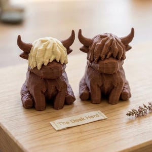 May include: Two brown Highland cow figurines with textured details and curved horns. One cow has a cream-colored fringe. A small tag with "The Desk Herd" is visible. The figurines are on a wooden surface.
