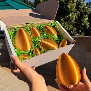 May include: A box of golden-colored, egg-shaped objects nestled in green decorative grass. One of the eggs is held in a person's hand. The box is white with a brown cardboard lid. The background includes green foliage and a blue sky.