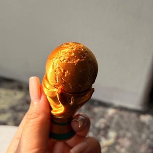 May include: A gold-colored trophy with a globe design held in a hand. The trophy has a detailed texture and a green base. The globe shows the continents. The trophy is a representation of a world championship.