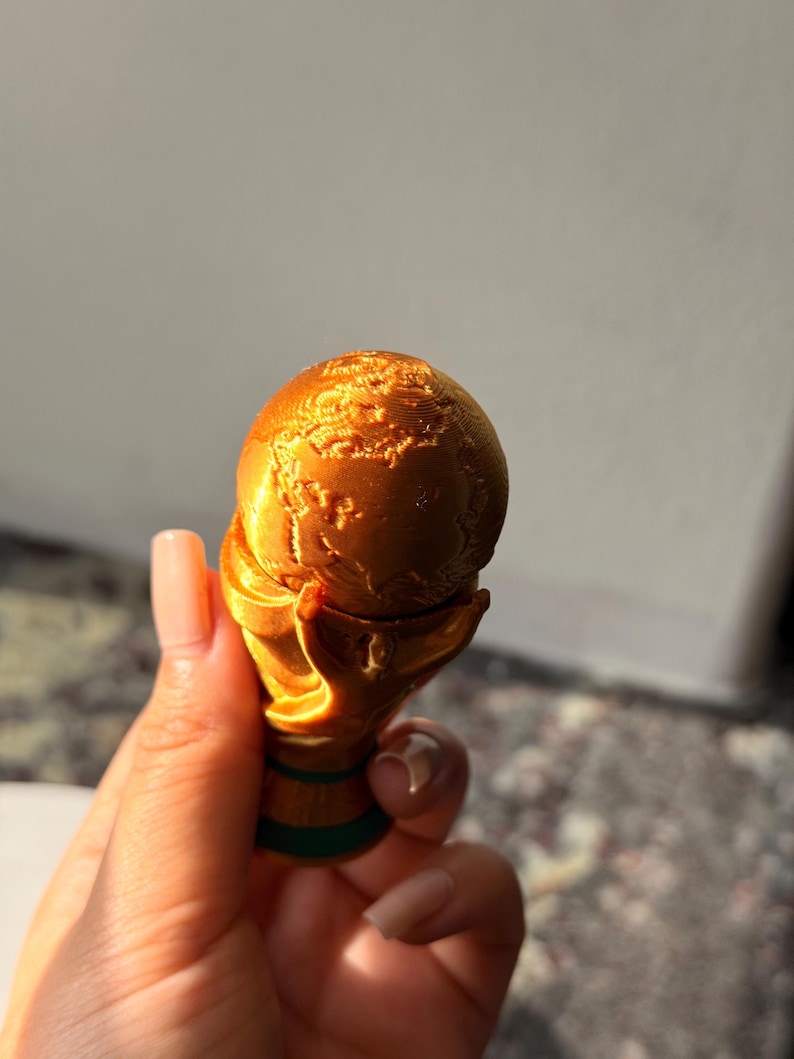 May include: A gold-colored trophy with a globe design held in a hand. The trophy has a detailed texture and a green base. The globe shows the continents. The trophy is a representation of a world championship.