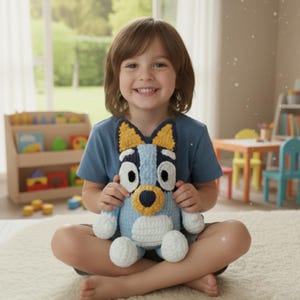 May include: A crocheted blue dog plush toy with yellow ears and a white belly is held by a child. The toy has large black and white eyes and a yellow nose. The child is wearing a blue shirt and sitting on a white rug.