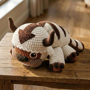 May include: A crocheted plush toy of a bison-like creature, featuring a cream-colored body with brown accents. The head has a brown arrow marking and two curved horns. The toy is resting on a wooden surface.