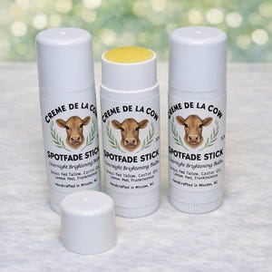 May include: Three white tubes of "Creme de la Cow" Spotfade Stick overnight brightening balm. The tubes have a cow graphic and text. One tube has a yellow balm exposed, and a cap is off. The product contains grass-fed tallow, castor oil, lemon peel, and frankincense.