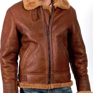 Può includere: Giacca in pelle marrone con colletto e fodera in montone. La giacca presenta una chiusura con cerniera completa, due tasche anteriori e finiture in montone sui polsini e sull'orlo. Indossata sopra una camicia bianca e jeans blu.