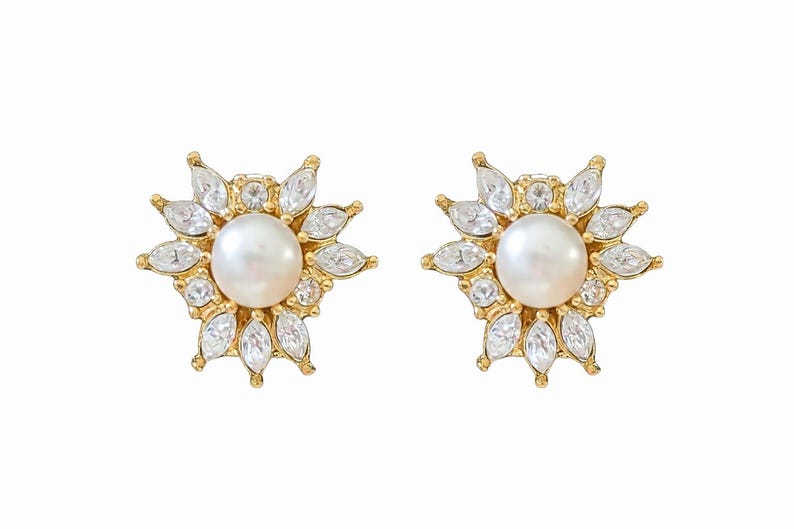May include: A pair of gold-toned stud earrings. Each earring features a central white pearl surrounded by clear, marquise-cut crystals, creating a floral or starburst design. The earrings are set against a white background.