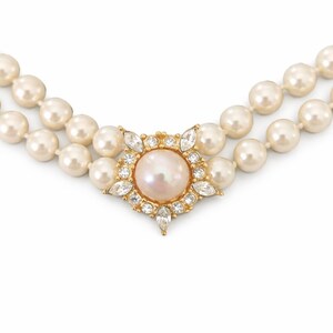 May include: A double-strand pearl necklace with a central pendant. The pendant features a large, light pink pearl surrounded by clear crystals and a gold-toned setting. The pearls are a creamy white color.