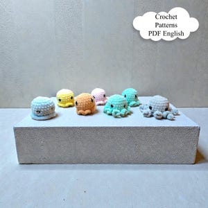 Könnte beinhalten: Eine Sammlung kleiner, farbenfroher gehäkelter Oktopus-Spielzeuge. Die Oktopusse sind in verschiedenen Farben erhältlich, darunter Blau, Gelb, Orange, Rosa und Grün. Der Text "Crochet Patterns PDF English" befindet sich in einer weißen Wolkenform.