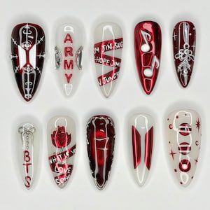 May include: A set of ten artificial nails with various designs. The nails feature a mix of red, white, and silver colors, with some adorned with text, musical notes, and other decorative elements. The nails are designed for cosmetic use.