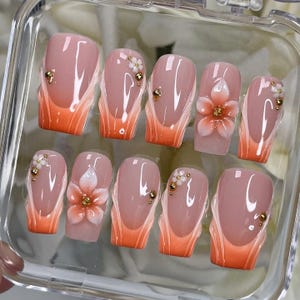 May include: A set of ten artificial nails with a French manicure design. The nails feature a nude base with an orange gradient at the tips. Some nails are adorned with small white flowers and gold accents, while others have gold beads.