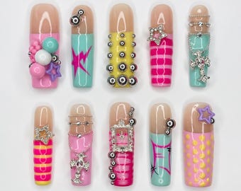 Puede incluir: Un juego de diez uñas postizas con diversos diseños en colores rosa, amarillo y turquesa. Los adornos incluyen tachuelas plateadas, cadenas metálicas, dijes de cruz, formas de estrella, patrones de rayas y un pequeño marco de fotos plateado.