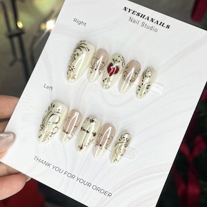 May include: A set of ten almond-shaped press-on nails on a white display card labeled NYESHANAILS Nail Studio. The nails feature a cream base with gold filigree swirls, small red dots, and a single prominent red heart design on one nail.