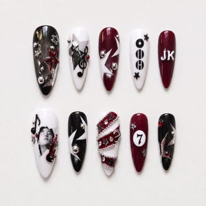 May include: A set of ten almond-shaped press-on nails featuring a color palette of black, white, and deep red. Designs include metallic star studs, a treble clef, the letters JK, a portrait, a belt-like strap, the number 7, and silver spheres.