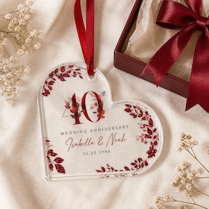 May include: A heart-shaped clear acrylic ornament featuring red glitter leaf accents, the text 40th WEDDING ANNIVERSARY, the names Isabella & Noah, and the date 11.25.1986. The ornament hangs from a red satin ribbon and is displayed near a gift box.