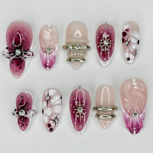 May include: A set of ten artificial nails in various designs. Some nails feature a gradient of pink and white, while others have intricate floral or star-like embellishments. Several nails are adorned with silver rings.