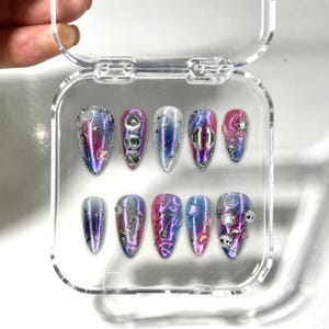 May include: A set of ten press-on nails in a clear plastic case, featuring a gradient of pink, purple, and blue. The nails are decorated with silver metallic accents, including the text BTS, the number 7, musical notes, stars, and small spheres.