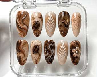 May include: A set of ten press-on nails in a clear plastic case. The nails feature various coffee-themed designs, including swirls of brown and tan, coffee beans, and latte art. Some nails have gold glitter accents.