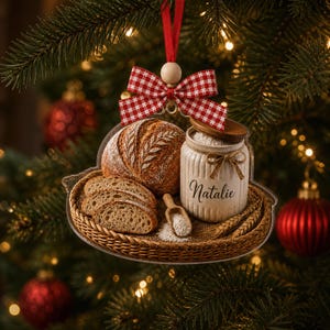 May include: A flat, clear acrylic Christmas ornament featuring a printed image of a round loaf of bread, three bread slices, and a beige ceramic jar labeled Natalie with a wooden lid and scoop, all resting on a woven tray with a red gingham bow.