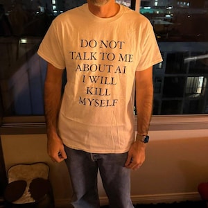 Könnte beinhalten: Cremefarbenes T-Shirt mit dem schwarzen Text "DO NOT TALK TO ME ABOUT AI I WILL KILL MYSELF". Das kurzärmelige Shirt scheint aus Baumwolle zu sein. Die Person trägt auch eine blaue Jeans.