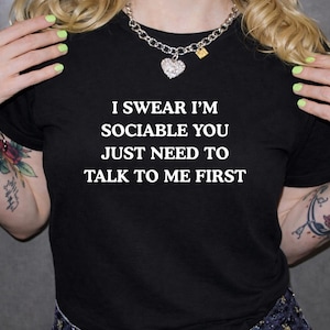 May include: Black t-shirt with the white text: "I SWEAR I'M SOCIABLE YOU JUST NEED TO TALK TO ME FIRST." The person is wearing a silver chain necklace with a heart pendant. The person has tattoos on their arms.