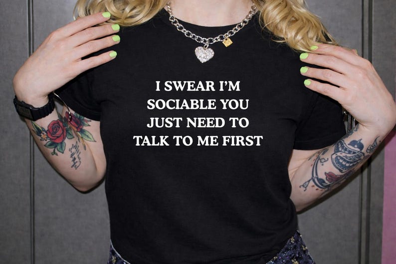 May include: Black t-shirt with the white text: "I SWEAR I'M SOCIABLE YOU JUST NEED TO TALK TO ME FIRST." The person is wearing a silver chain necklace with a heart pendant. The person has tattoos on their arms.