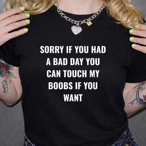 May include: Black t-shirt with white text that reads "SORRY IF YOU HAD A BAD DAY YOU CAN TOUCH MY BOOBS IF YOU WANT." The person is wearing a silver necklace and has tattoos on their arms.