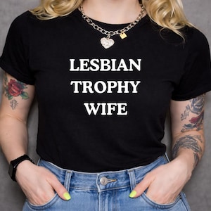 Könnte beinhalten: Schwarzes T-Shirt mit dem weißen Text "LESBIAN TROPHY WIFE". Die Person trägt eine silberne Halskette mit Herzanhänger und blaue Jeans. Die Person hat Tätowierungen auf den Armen.