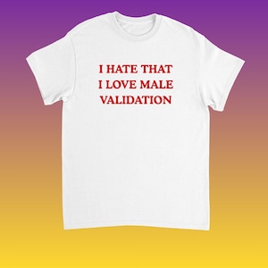 May include: White t-shirt with the red text "I HATE THAT I LOVE MALE VALIDATION". The shirt is made of a soft material and has a crew neck. The background is a gradient of purple, orange, and yellow.