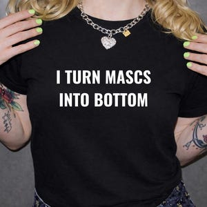 May include: Black t-shirt with the white text "I TURN MASCS INTO BOTTOM". The shirt is short-sleeved. The person is wearing a silver chain necklace with a heart pendant. The person has tattoos on their arms.