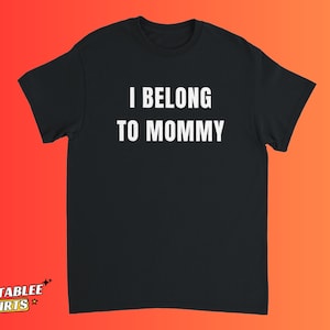 May include: Black t-shirt with the white text "I BELONG TO MOMMY". The shirt has short sleeves and a crew neck. The text is centered on the front of the shirt. The background is a gradient of orange and red.