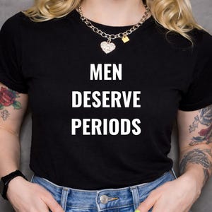 Könnte beinhalten: Schwarzes T-Shirt mit dem weißen Text "MEN DESERVE PERIODS". Die Person trägt eine silberne Halskette mit Herzanhänger und eine schwarze Uhr. Die Person trägt auch eine blaue Jeans.