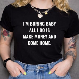 Könnte beinhalten: Ein schwarzes Kurzarm-T-Shirt mit dem weißen Aufdruck: I'M BORING BABY ALL I DO IS MAKE MONEY AND COME HOME. Das T-Shirt wird mit einer blauen Jeans kombiniert und dazu eine silberfarbene Kette mit einem herzförmigen Anhänger getragen.