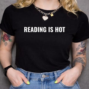 Könnte beinhalten: Schwarzes T-Shirt mit dem weißen Text "READING IS HOT". Das Shirt wird mit einer blauen Jeans getragen. Die Person trägt eine silberne Halskette mit Herzanhänger und eine schwarze Uhr.