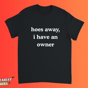 Może przedstawiać: Czarny t-shirt z białym napisem "hoes away, i have an owner". Koszulka ma krótkie rękawy i okrągły dekolt. Koszulka jest prezentowana na tle gradientu pomarańczowo-czerwonego.
