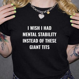 Könnte beinhalten: Schwarzes T-Shirt mit dem weißen Text "I WISH I HAD MENTAL STABILITY INSTEAD OF THESE GIANT TITS." Die Person trägt eine silberne Halskette mit Herzanhänger. Die Person hat Tätowierungen auf den Armen.