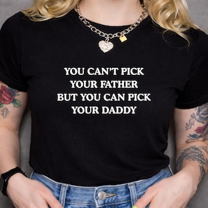 May include: Black t-shirt with the white text: "YOU CAN'T PICK YOUR FATHER BUT YOU CAN PICK YOUR DADDY." The shirt is worn with blue jeans and a silver necklace with a heart pendant.