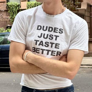 Puede incluir: Camiseta blanca con el texto "DUDES JUST TASTE BETTER" en letras negras. La camiseta se combina con vaqueros de mezclilla azul oscuro. Se ven una cadena y una pulsera plateadas.