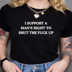Puede incluir: Camiseta negra con el texto blanco "I SUPPORT A MAN'S RIGHT TO SHUT THE FUCK UP". La camiseta se lleva con vaqueros azules. La persona tiene tatuajes en los brazos y lleva un reloj y un collar.