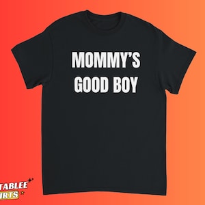 Może przedstawiać: Czarny t-shirt z krótkim rękawem z białym napisem "MOMMY'S GOOD BOY" wydrukowanym pogrubioną, bezszeryfową czcionką na klatce piersiowej. Odzież posiada okrągły dekolt i standardowy krój, prezentując prosty design o wysokim kontraście.
