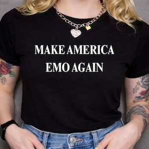 May include: Black t-shirt with the white text "MAKE AMERICA EMO AGAIN." The shirt is a crew neck style. The person is wearing a silver chain necklace with a heart pendant and a black watch.