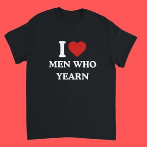 Op de afbeelding: Zwart t-shirt met de tekst "I ❤️ MEN WHO YEARN" in wit en rood. Het shirt heeft een ronde hals en korte mouwen. De achtergrond is effen rood.