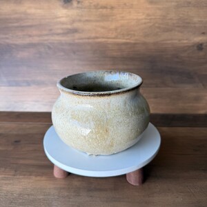 May include: A small, round, beige ceramic pot with a dark brown rim, sitting on a white, round, wooden stand with three brown legs. The pot has a textured surface and a slightly glossy finish. The background is a wooden surface.