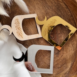 Könnte beinhalten: Dekorative Serviettenringe aus Holz, Gold und Silber. Jeder Ring hat ein Mond-Design und arabische Kalligraphie. Die Ringe liegen auf einer Holzoberfläche mit einer weißen Serviette.