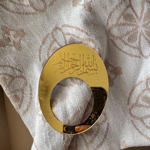 Könnte beinhalten: Ein goldfarbener Serviettenring mit arabischer Schrift liegt auf einer gefalteten beigefarbenen Serviette mit floralem Muster. Der Serviettenring hat eine Halbmondform und ist reflektierend.