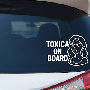 May include: White vinyl car decal with the text "TOXICA ON BOARD" and a line drawing of a woman with a stern expression. The decal is designed to be applied to a car window or bumper.